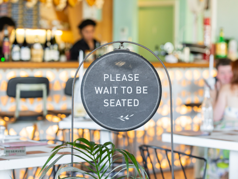 A restaurant sign that reads 'Please wait to be seated' with a plant in the foreground, and a blurred background showing a bar area and a person sitting at a table.