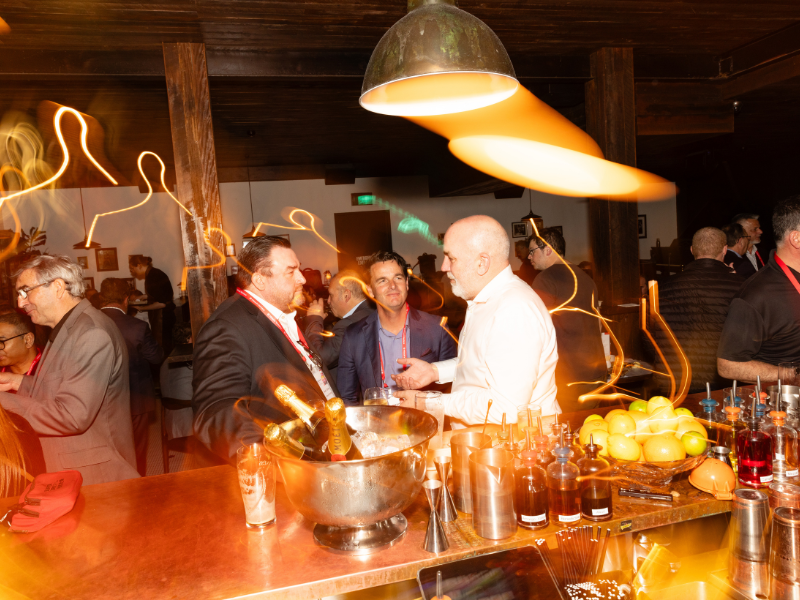 People socializing at a bar with bottles of alcohol and apples on the counter, warm lighting, and a wooden ceiling.
