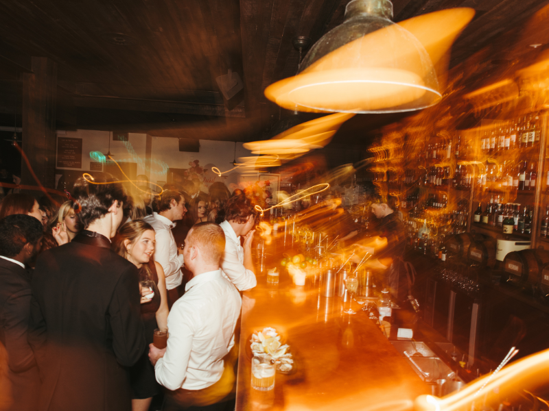 People gathering at a bar with warm lighting, busy atmosphere, and blurred light trails from motion.