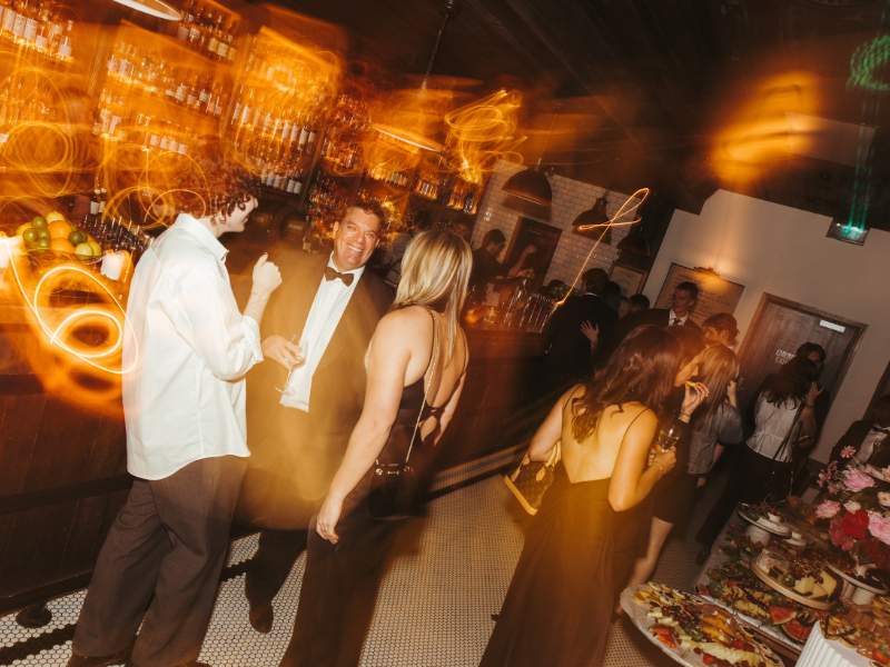 People dancing and socializing in a lively, dimly-lit bar or club with a bar counter and food tables.