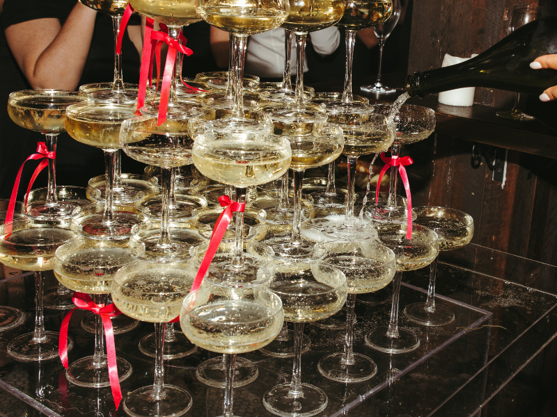 Multiple champagne glasses filled with champagne, some with red ribbons tied around the stems, stacked in a pyramid formation at a celebration or party.