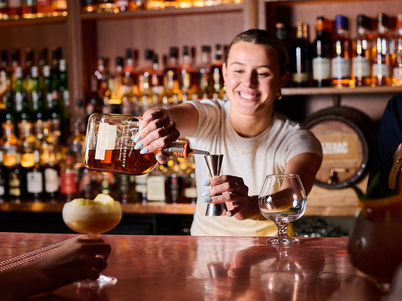 A woman bartends at a bar, pouring a drink from a bottle into a jigger, with shelves of liquor bottles in the background.