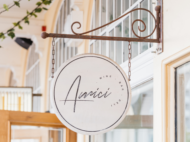 Round white sign hanging from a wrought iron bracket with the word 'Avaici' in elegant script, and the words 'Wine', 'Bar', and 'Deli' around the edge.