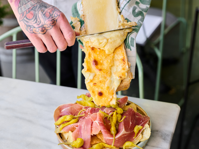 Person with tattooed hand serving pizza with melted cheese, ham, and pickled peppers on a marble table.