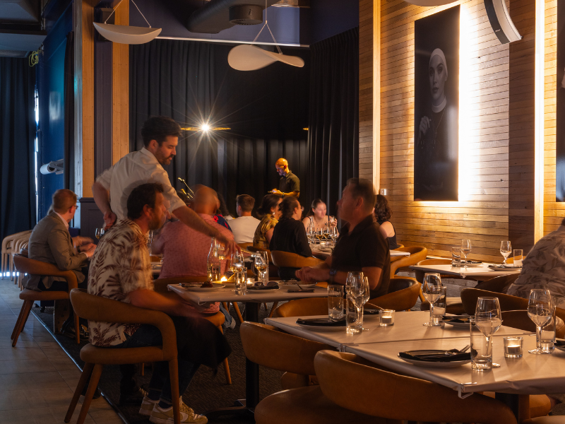 A lively restaurant scene with diners seated at tables, enjoying meals and drinks. A server is pouring a drink, and a musician is performing on stage in the background. The decor includes wooden wall panels and a large black-and-white portrait on the