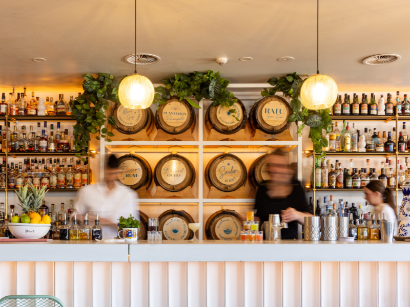 Bar with bottles of alcohol on shelves, decorative barrels on the wall with greenery, and two pendant lights hanging from the ceiling.