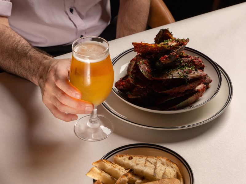 Person holding a glass of beer next to a plate of cooked ribs garnished with herbs, with a side of bread on the table.