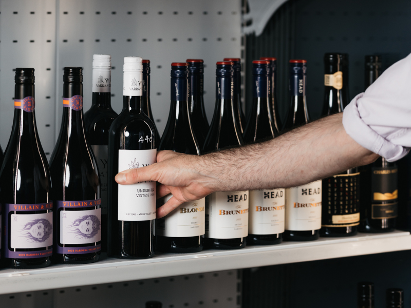 A person with a light-colored sleeve reaching for a bottle of wine on a store shelf. The shelf contains various bottles of red wine with different labels and caps.