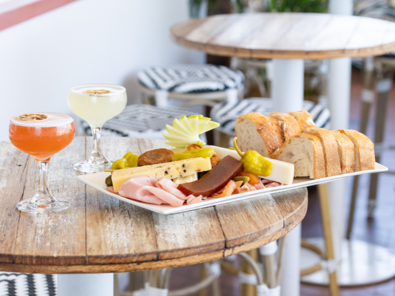 Charcuterie board with cheeses, meats, and bread, two cocktails with lemon slices garnishes on a rustic wooden table in a cozy indoor setting