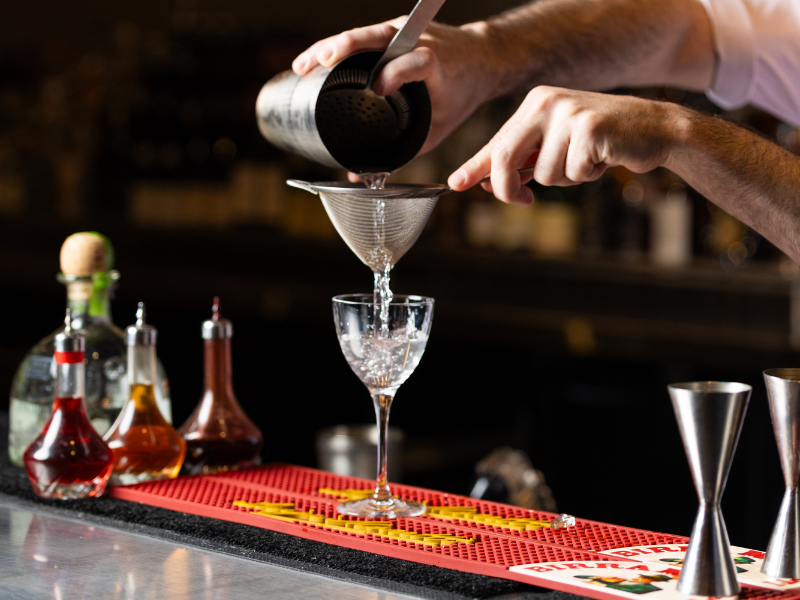 Bartender pouring a liquid through a fine mesh strainer into a cocktail glass at a bar counter with various bottles of liquor in the background.