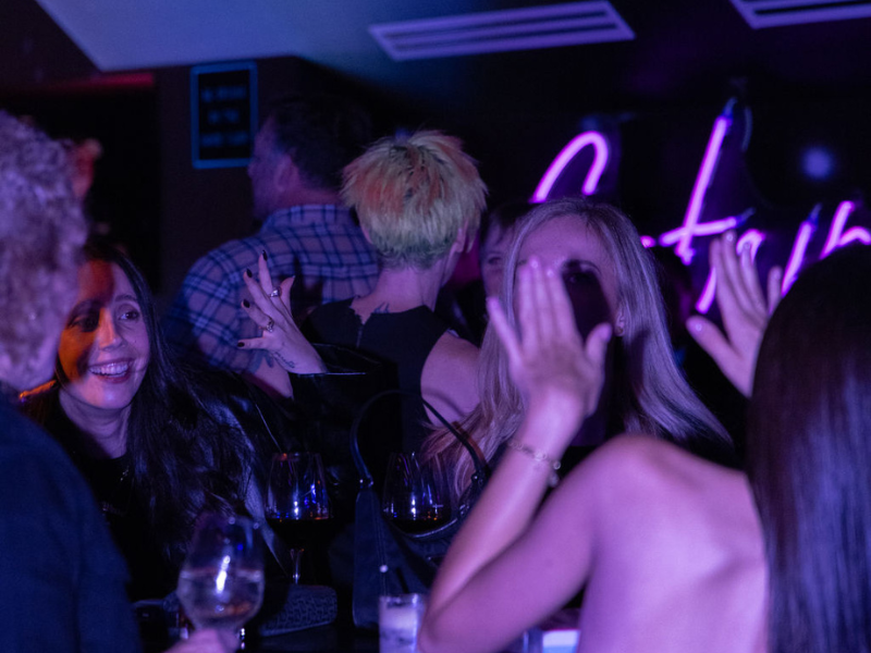 Group of people socializing and having drinks at a bar or nightclub with purple neon lighting.