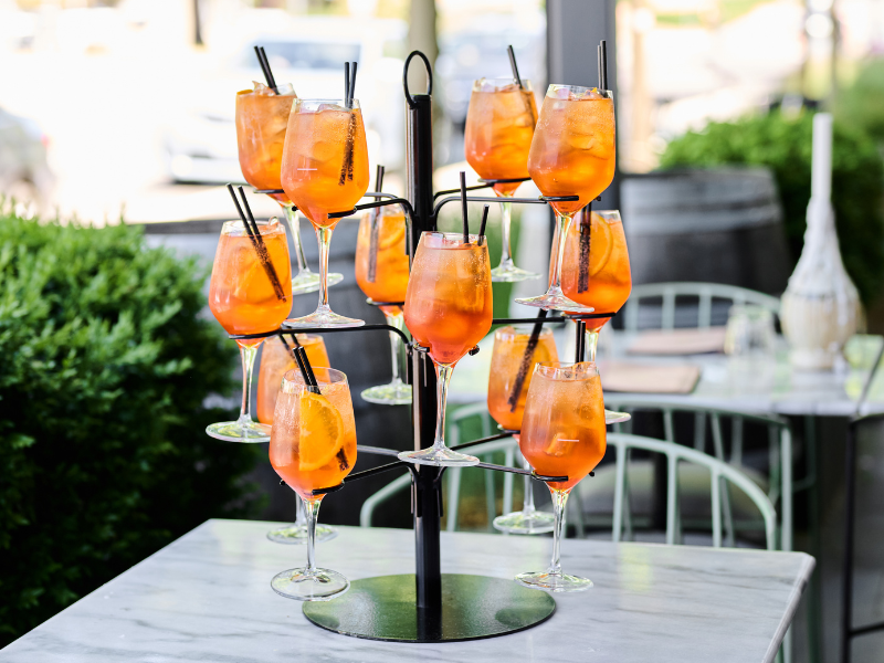A tiered stand with multiple glasses of orange-colored cocktails with ice, garnished with slices of orange and black straws, placed on a white marble table outdoors.