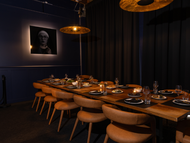 Empty dining table set with black placemats, water glasses, small plates of food, and candles in a dimly lit room with blue walls and black curtains, featuring a black-and-white portrait of a woman on the wall.