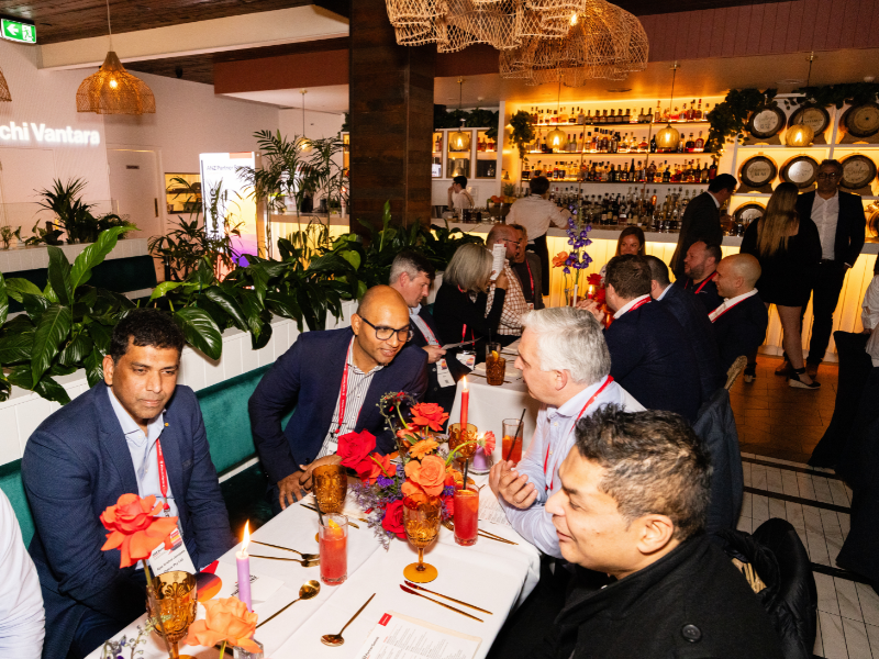 People sitting around a decorated table with flowers and candles at a restaurant or event, with a bar and other guests in the background.