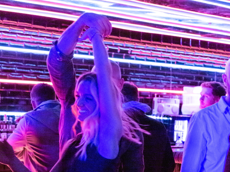 A woman is dancing with her arms raised in a bar or nightclub with neon lighting and other people in the background.