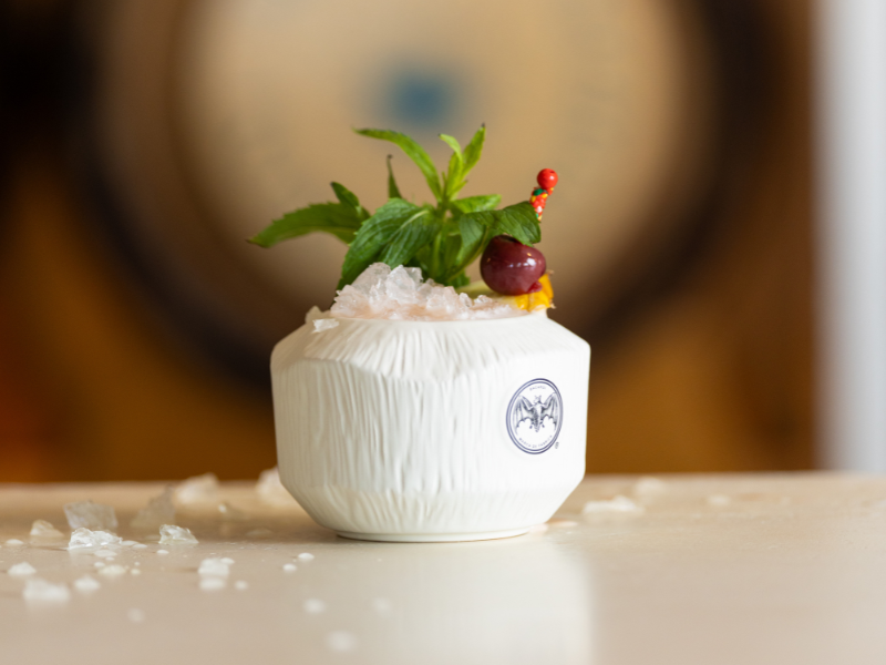 Decorative white ceramic container with a bat emblem, filled with ice, garnished with mint leaves, a cherry, and berries.