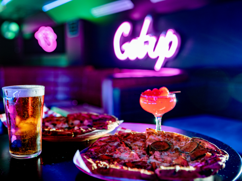 A bar scene with two pizzas, a glass of beer, and a cocktail with cherries, under neon lighting and a pink neon sign in the background.