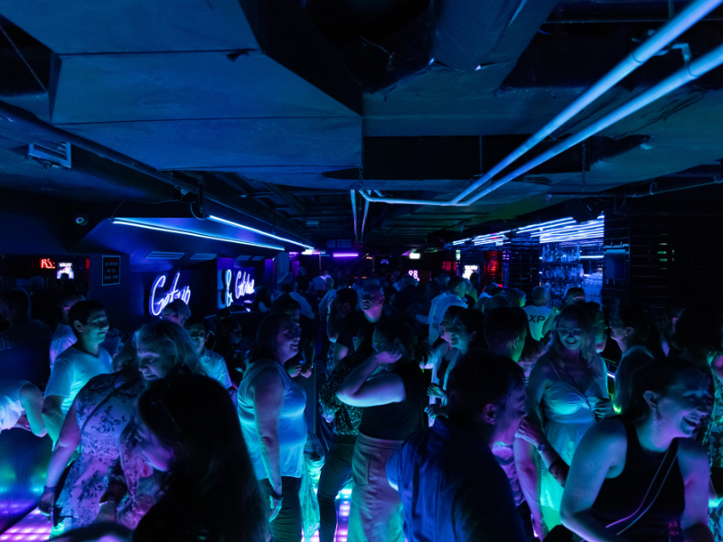 Crowd of people dancing and socializing at a nightclub with neon lighting and illuminated signs.