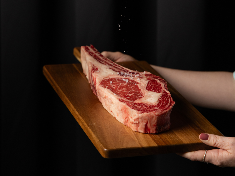 Person seasoning a large raw steak on a wooden cutting board.