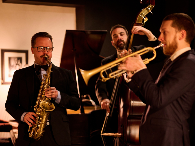 Three musicians playing jazz instruments: a man with glasses and a tuxedo playing a saxophone, a man with a beard playing a double bass, and a man with a beard playing a trumpet, in a dimly lit performance setting.