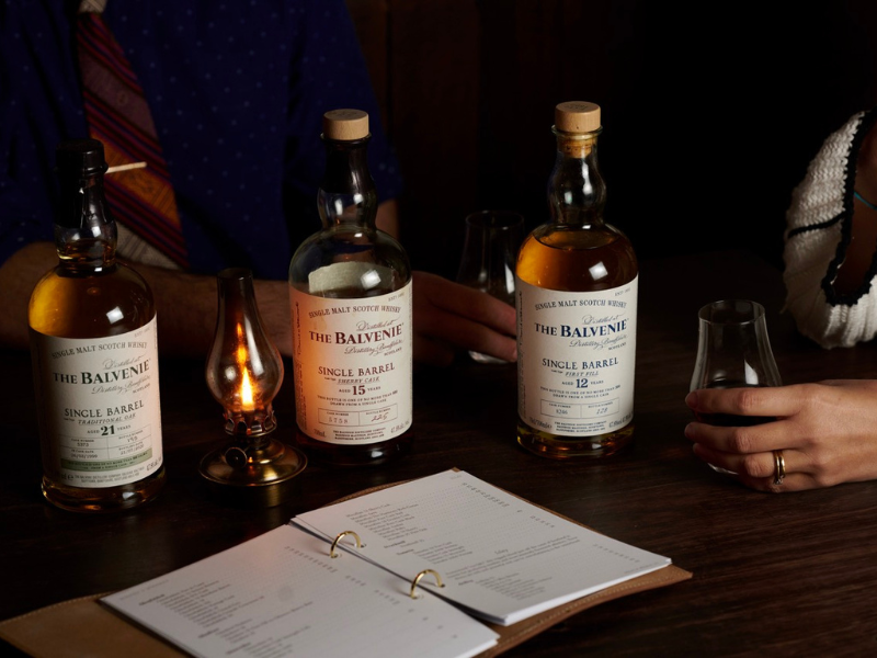 Three bottles of The Balvenie single malt Scotch whisky on a dark wooden table, with a small oil lamp, a tasting guide, and two people holding glasses.