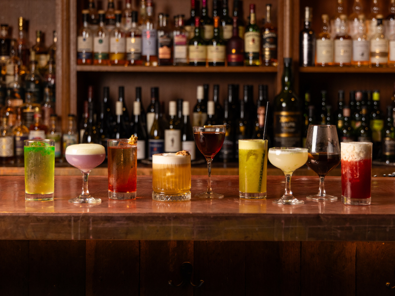 A row of colorful cocktails on a wooden bar counter with a variety of liquor bottles in the background.