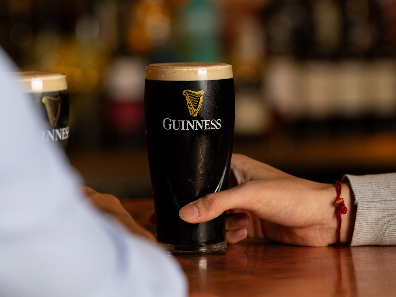 A person holding a pint of Guinness stout beer with a foamy head at a bar.