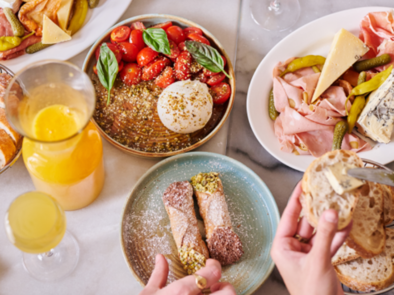 Various plates of food on a table, including a bowl of cherry tomatoes with basil, a plate of charcuterie with cheese and pickles, a plate with cannoli, and glasses of orange juice.