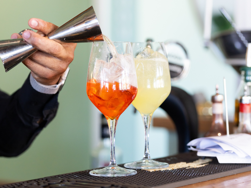 Bartender pouring a colorful cocktail over ice into a wine glass, with another yellow drink nearby, on a bar counter.