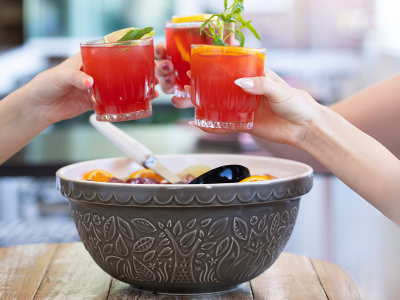 Three people raising glasses of red cocktails with garnishes over a bowl of fruit on a wooden table.