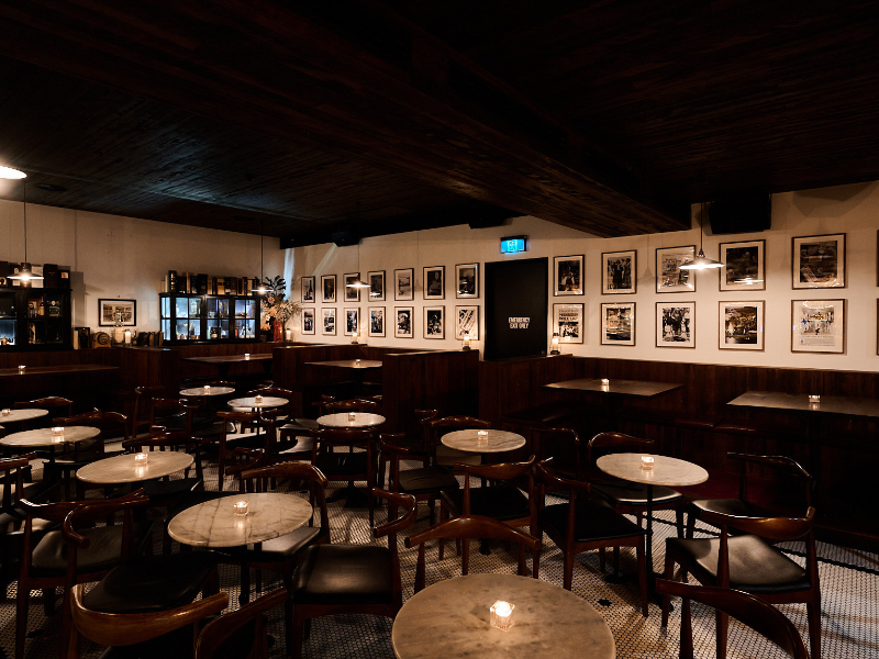 Empty restaurant with round marble tables, wooden chairs, wall decorated with black-and-white photos, dim lighting, and a dark wood ceiling.