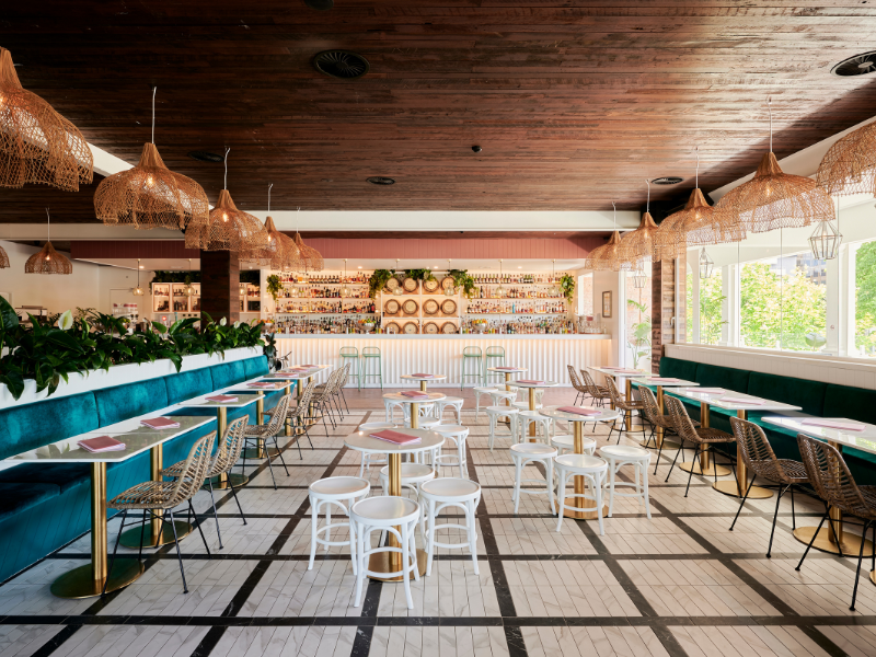 Interior of a modern restaurant or cafe with a wooden ceiling, hanging wicker pendant lights, and large windows. There are teal upholstered benches, white and wicker chairs, tables set with pink napkins, and a bar with bottles and decorative plants in the background.