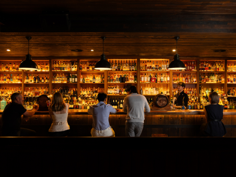 People sitting at a bar counter with liquor bottles behind the bar and a bartender serving drinks.