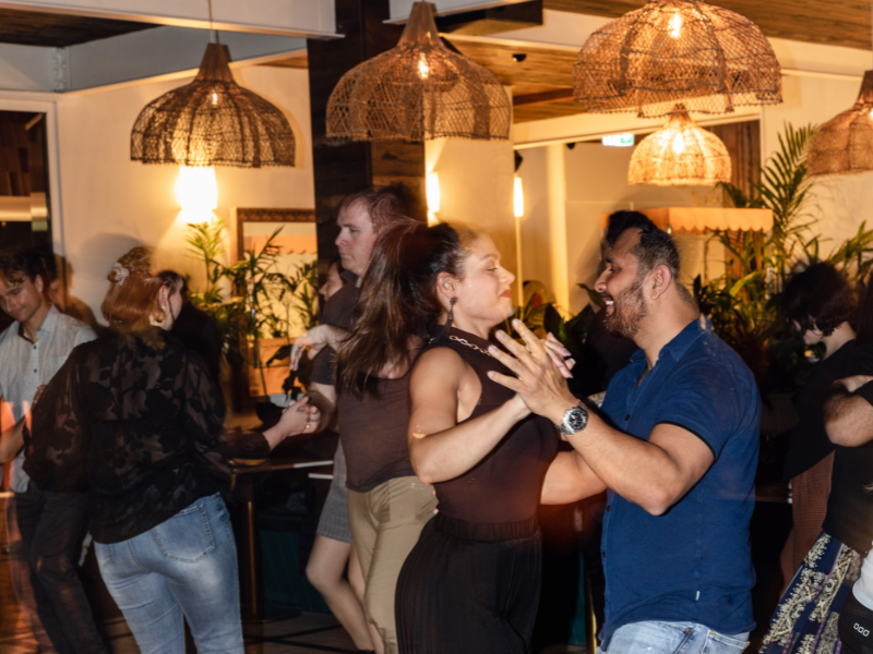 Couple dancing together at a social gathering in a warmly lit room with hanging wicker lamps and potted plants.