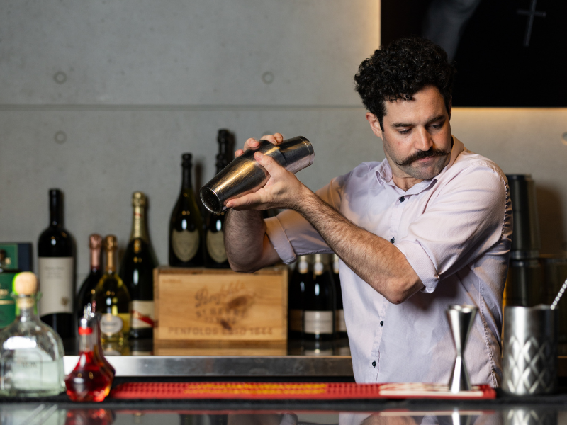 A bartender with dark curly hair and a mustache, wearing a light pink shirt, shakes a cocktail shaker behind a bar counter with various bottles visible in the background.