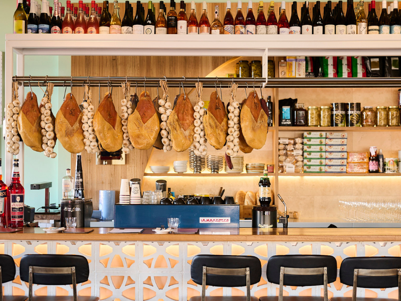 A bar or restaurant counter with hanging cured ham legs and garlic bulbs, bottles of wine on a shelf above, and various glassware, cans, and food items on the shelves behind the counter.