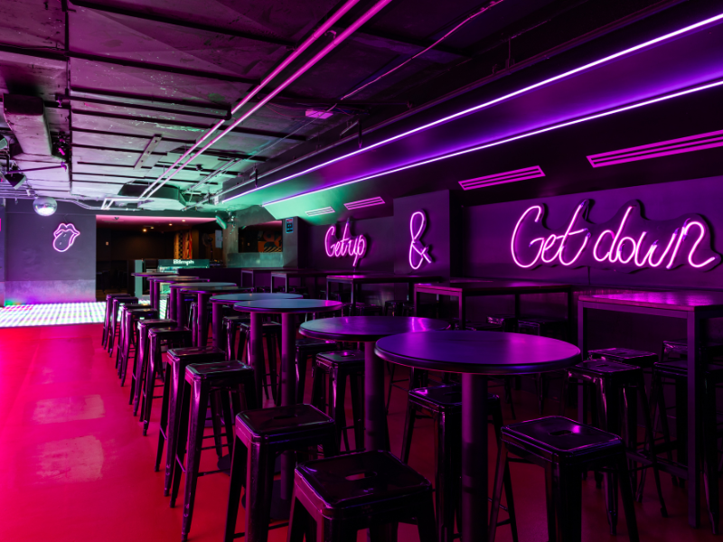 88mph Canberra bar interior with neon purple signs saying 'Get up & Get down,' and a neon lips sign, with a row of high-top tables and stools under purple and green light.
