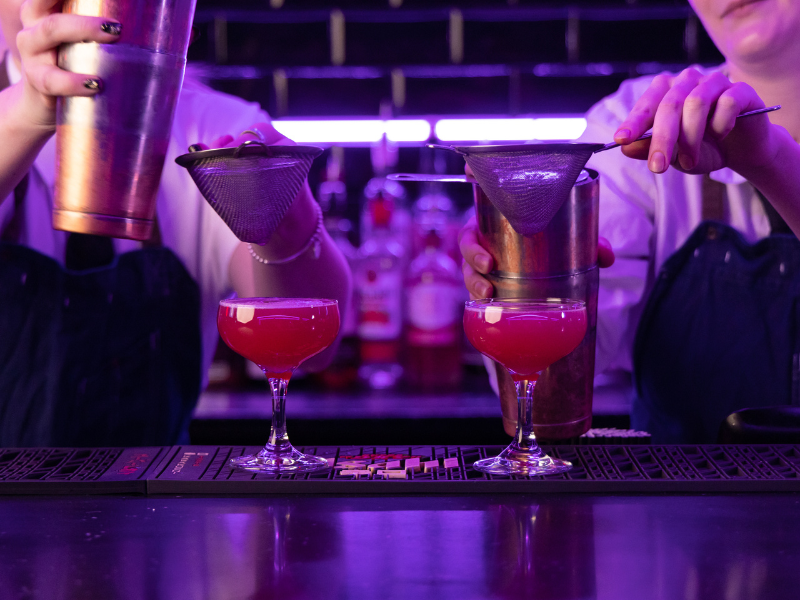 Two bartenders preparing cocktails with strainer, with pink cocktails in coupe glasses on bar counter, dimly lit bar with bottles in background.