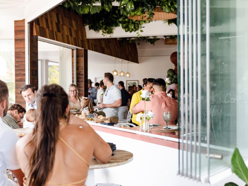 People socializing and dining inside a modern, well-lit restaurant with greenery and wood accents.
