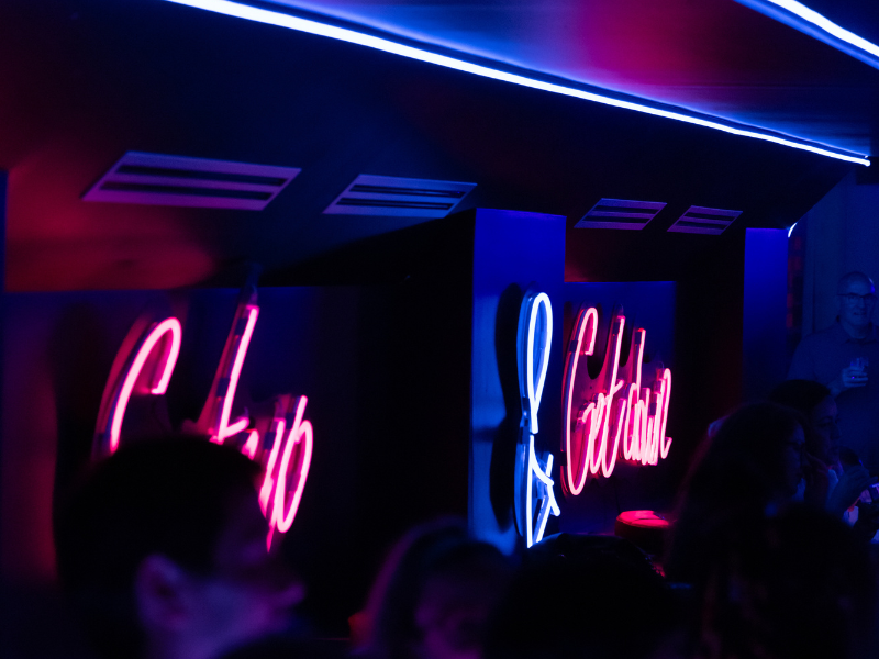 Neon signs reading 'Club' and 'Cacciator' in a dark, colorful nightclub setting.