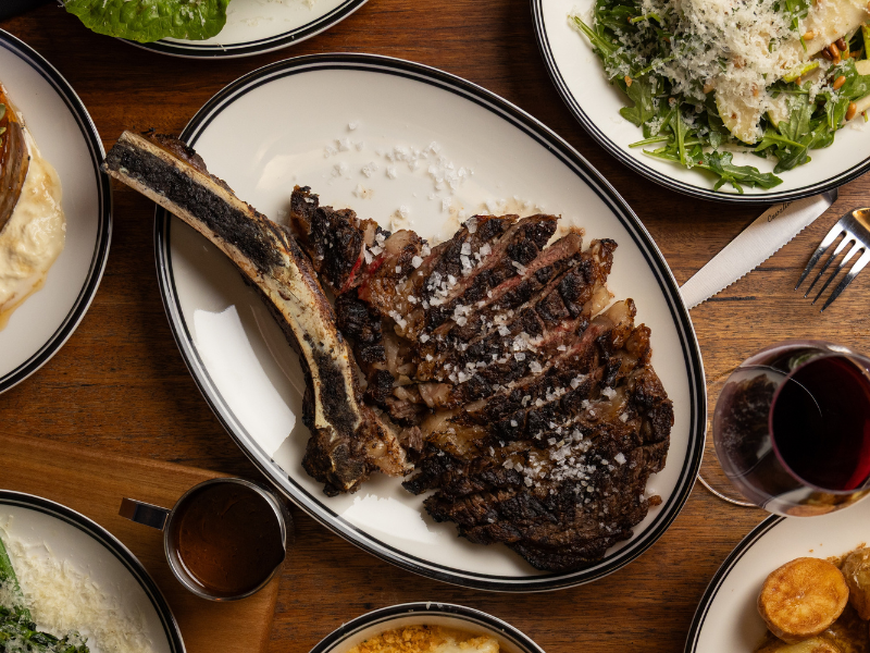 A grilled steak with a bone on a white oval plate, topped with grated cheese, surrounded by other plates of salad, wine, and side dishes on a wooden table.