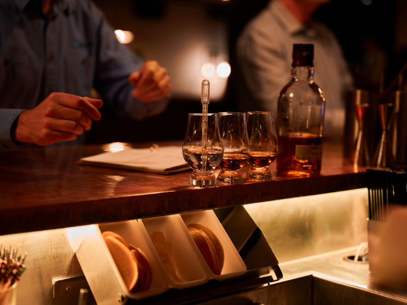 Bar scene with three glasses of whiskey, a bottle of whiskey, and a stir stick on the counter, with hands of bartender and customer visible.