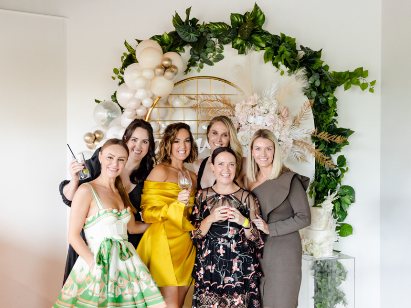 Six women at a celebration, standing in front of a decorated backdrop with greenery and balloons, smiling and holding drinks.