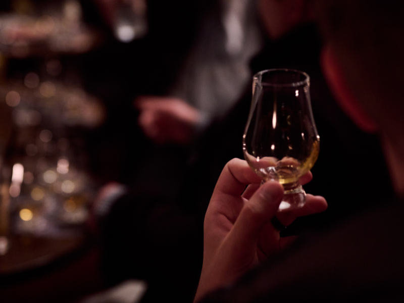 Person holding a glass of drink in a dimly lit bar or lounge, with blurred background of other people and glasses.