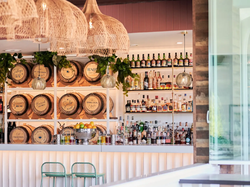 Bar with liquor bottles, wooden barrels, and hanging wicker lamps in a bright, modern setting.