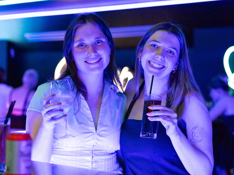 Two women smiling at a bar, holding drinks, illuminated by colorful neon lights.