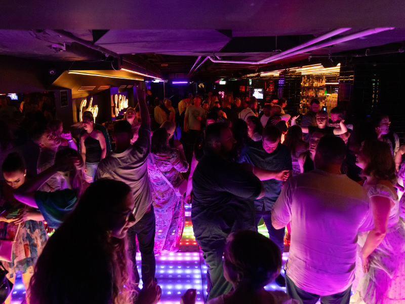 People dancing and socializing on a brightly lit dance floor at a nightclub with colorful lighting and a bar in the background.