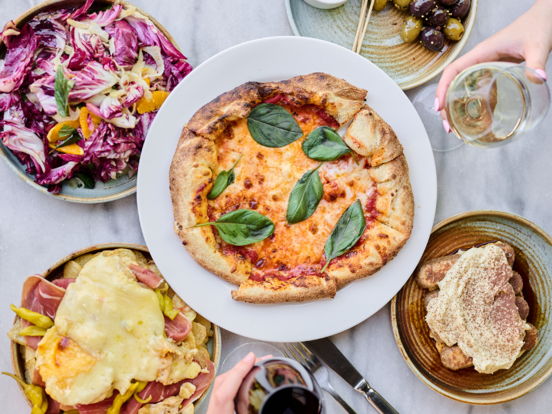 Flat lay of a pizza with basil leaves, surrounded by a salad, a dish with melted cheese and meats, a dish with breaded meat, and two glasses of white and red wine.