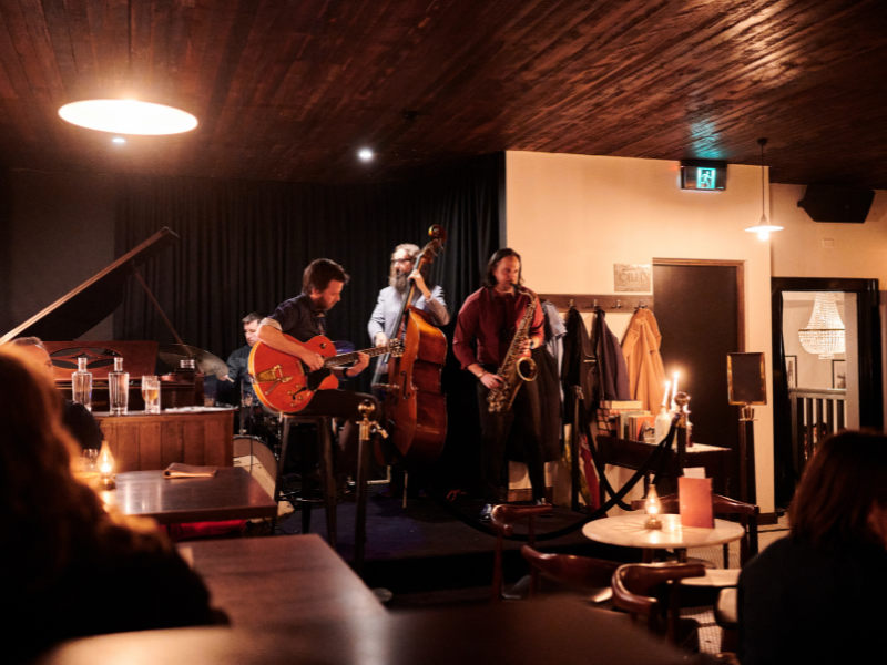Four musicians performing on a small stage in a cozy, dimly lit venue. One plays guitar, another on double bass, one on saxophone, and another on drums. Audience members sit at tables, some with drinks and candles.