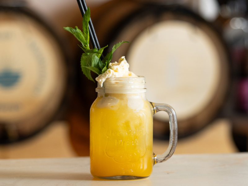 Mango lemonade in a glass jar with a handle, topped with whipped cream, a sprig of mint, and black straws, with wine barrels in the background.